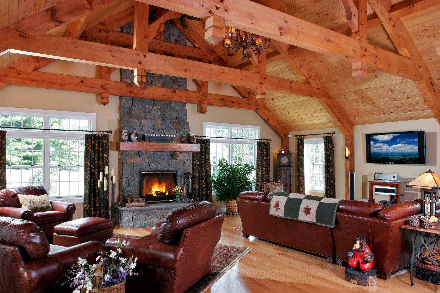 Teakwood Builders designed this great room to resemble a rustic Adirondack lodge, with abundant natural materials and a Rumford fireplace. A bluestone slab tops the raised hearth, while 4-5” thick split-stone veneer surrounds the fireplace and is carried to the ceiling. A rustic-style beam serving as the mantle complements the new timber trusses and posts.