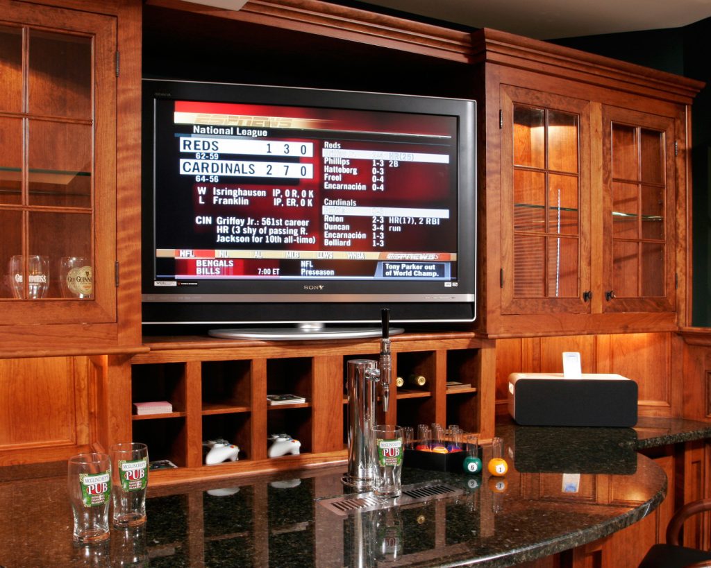 Teakwood designed and built this multi-function bar area. Custom cherry cabinets boast oil-rubbed bronze hardware, upper glass panels and discreet undercabinet lighting concealed by valance trim. Wine storage fits neatly beneath the television cabinet. Cut out of the granite countertop is a beer tap for the kegerator installed below.