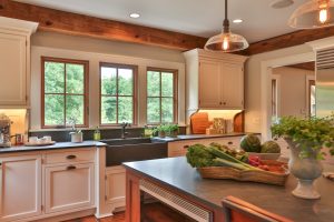 The barn kitchen renovation features a large apron sink, custom white cabinetry, a dark counter top and wooden windows. Kitchen design by Teakwood Builders, Saratoga Springs, NY.